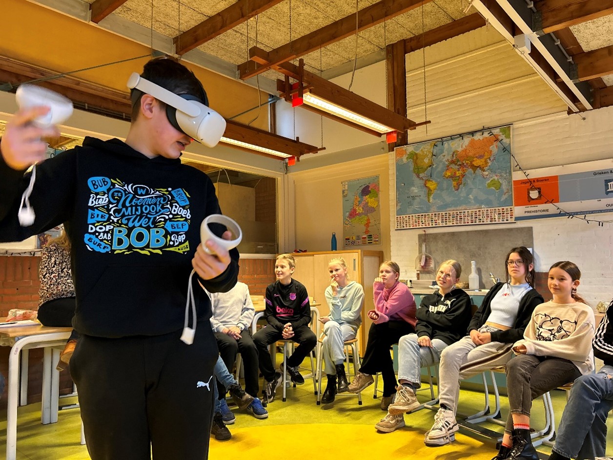 In een klaslokaal zitten leerlingen in een kring. Ze kijken naar een leerling op de voorgrond met een VR-bril op en controllers in zijn handen.