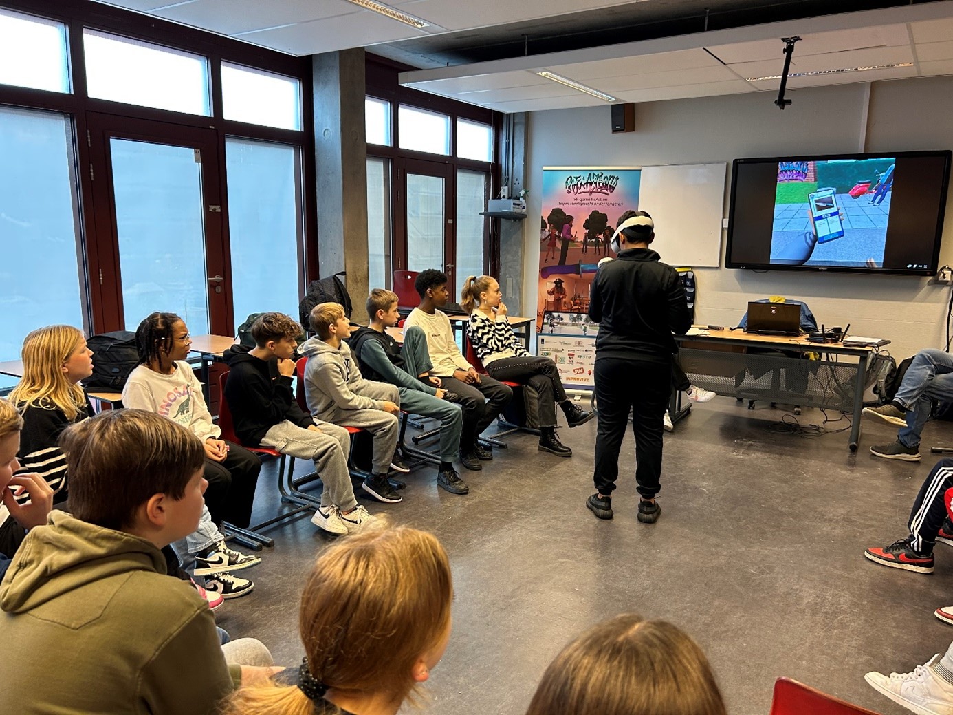 Leerlingen zitten in een kring en kijken hoe één leerling met een VR-bril op een spel speelt. Op de achtergrond is de game te zien op een smartboard.