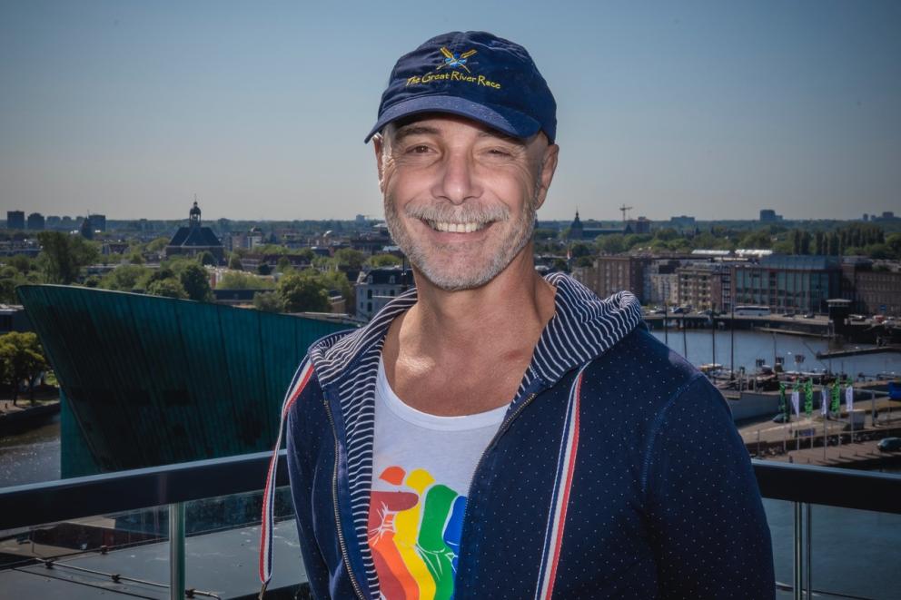 John Avis draagt een pet met The Great River Race en draagt een regenboog t-shirt, staat buiten met mooi weer bij Oosterdok
