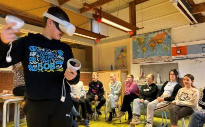 In een klaslokaal zitten leerlingen in een kring. Ze kijken naar een leerling op de voorgrond met een VR-bril op en controllers in zijn handen.