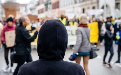 Foto van een protest. Op de voorgrond zie je iemand van achteren met een capuchon van diens hoodie omhoog.