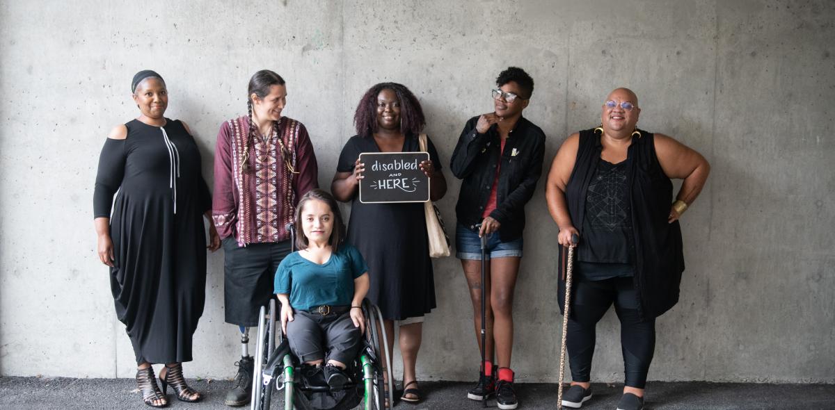 Groep van 6 personen met een beperking, waarvan één met een bordje 'Disabled and here'