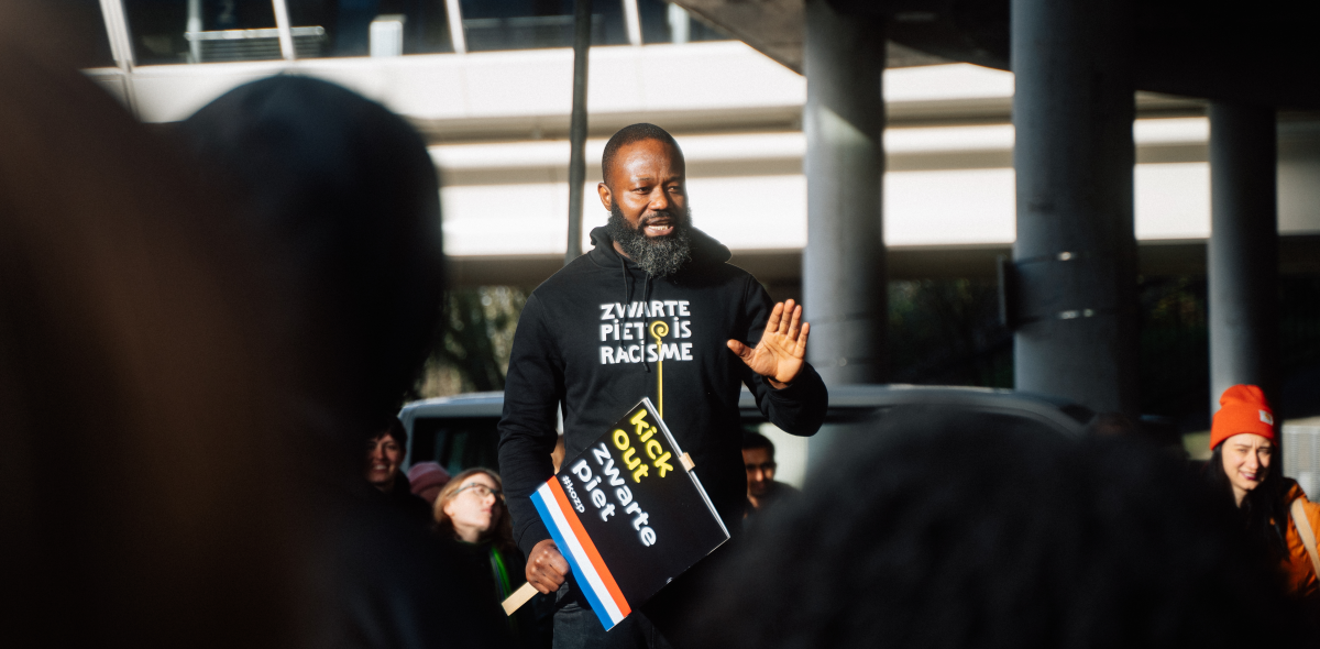Jerry Afriyie staat voor een menigte tijdens een demonstratie met een bord 'Kick out Zwarte Piet'