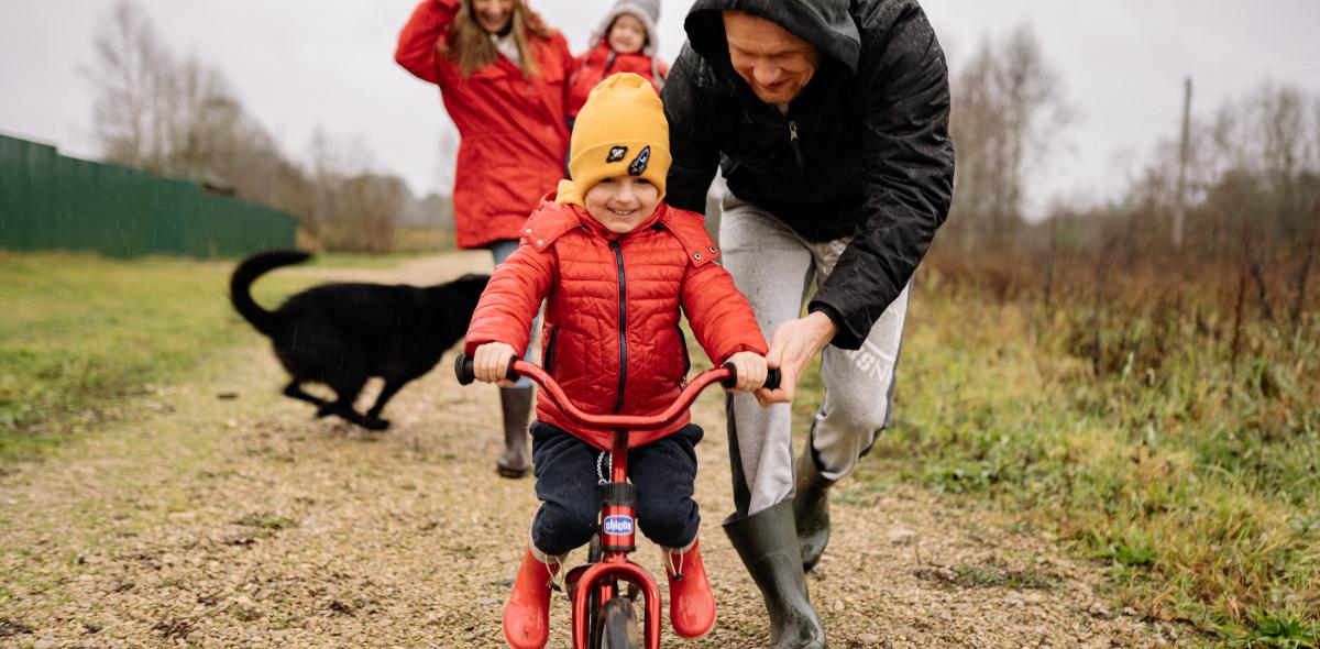 Een vader leert zijn kind fietsen. Op de achtergrond zijn ook de moeder met een kleiner kind op de arm en een hond te zien.